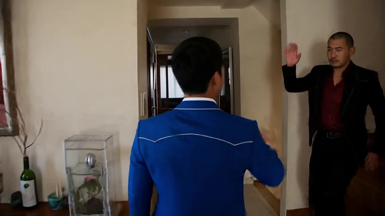 階段側で手を上げる黒いスーツと赤系のシャツの人物を青いスーツの主人公が迎える